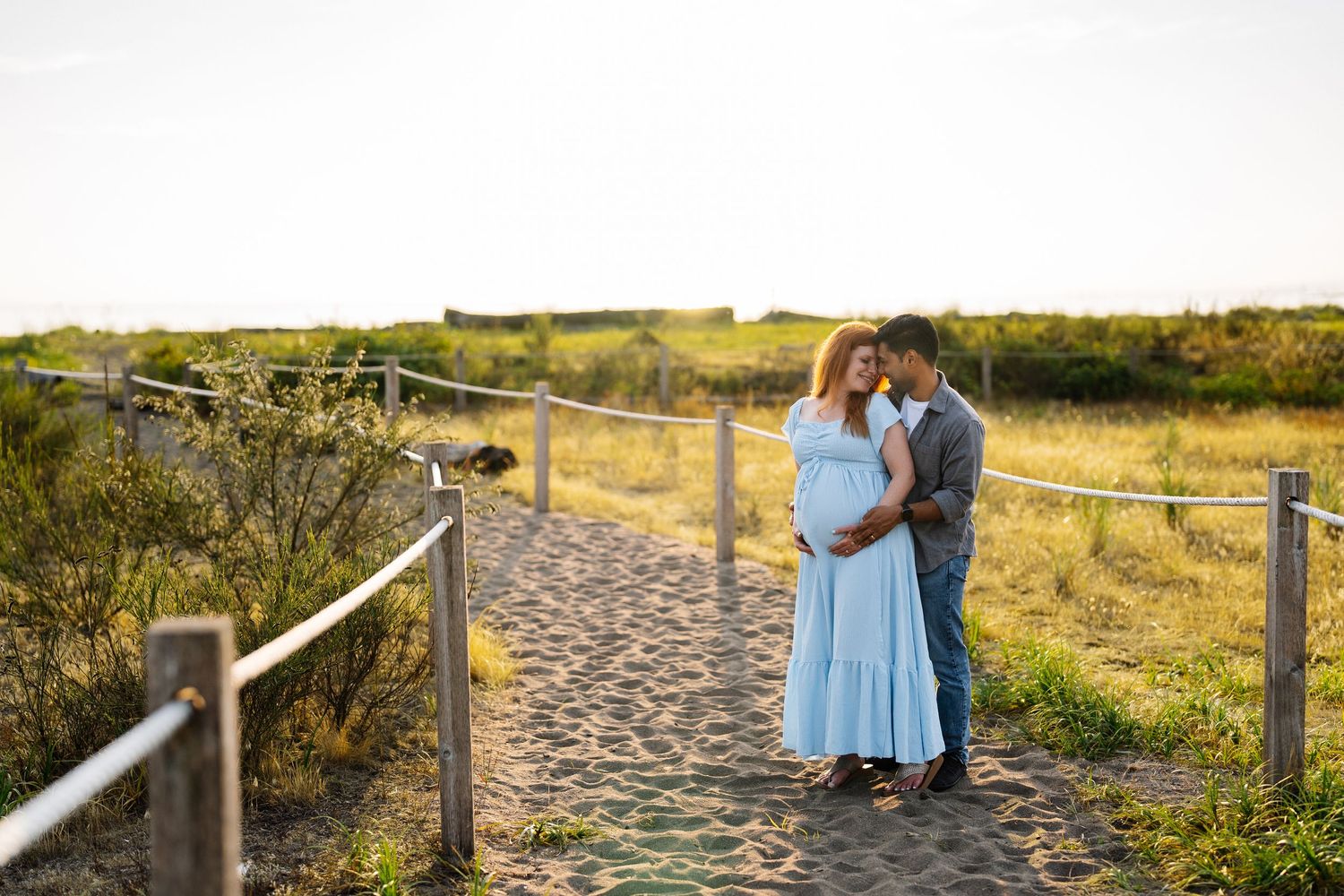 Expectant couple embracing on a sandy beach path lined with rope fencing at golden hour, woman in a flowing light blue maternity dress.