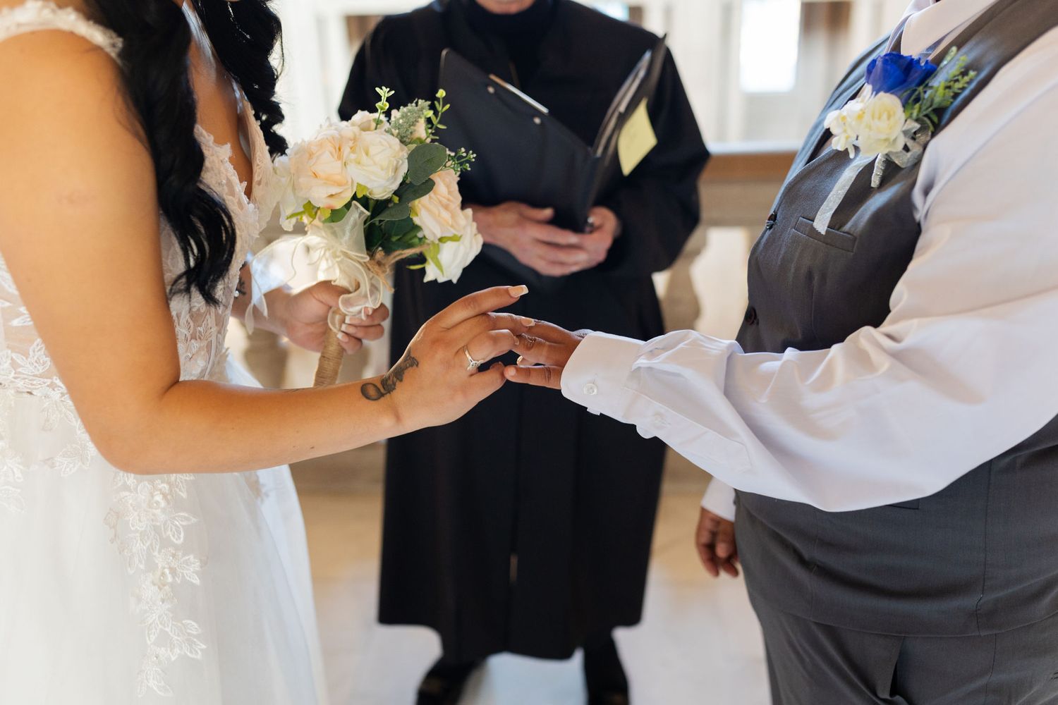 Bride placing wedding ring on groom's finger during ceremony, officiant holding book in background.