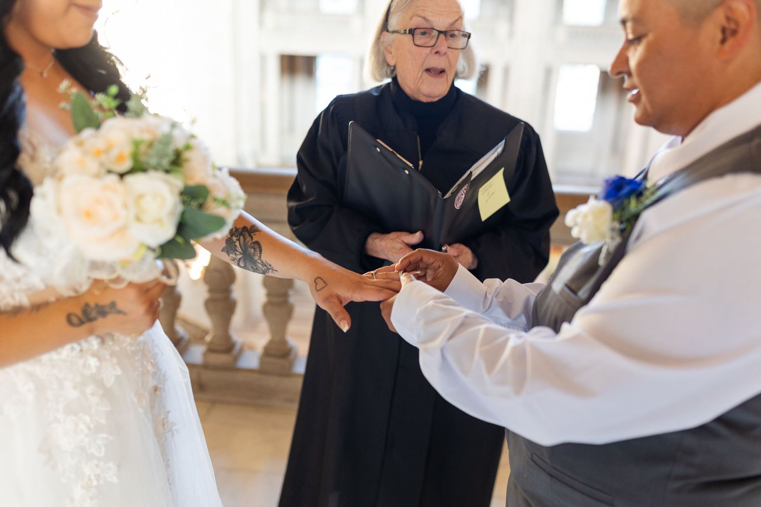 Officiant presiding over wedding ring exchange ceremony between bride holding bouquet and groom in formal attire.