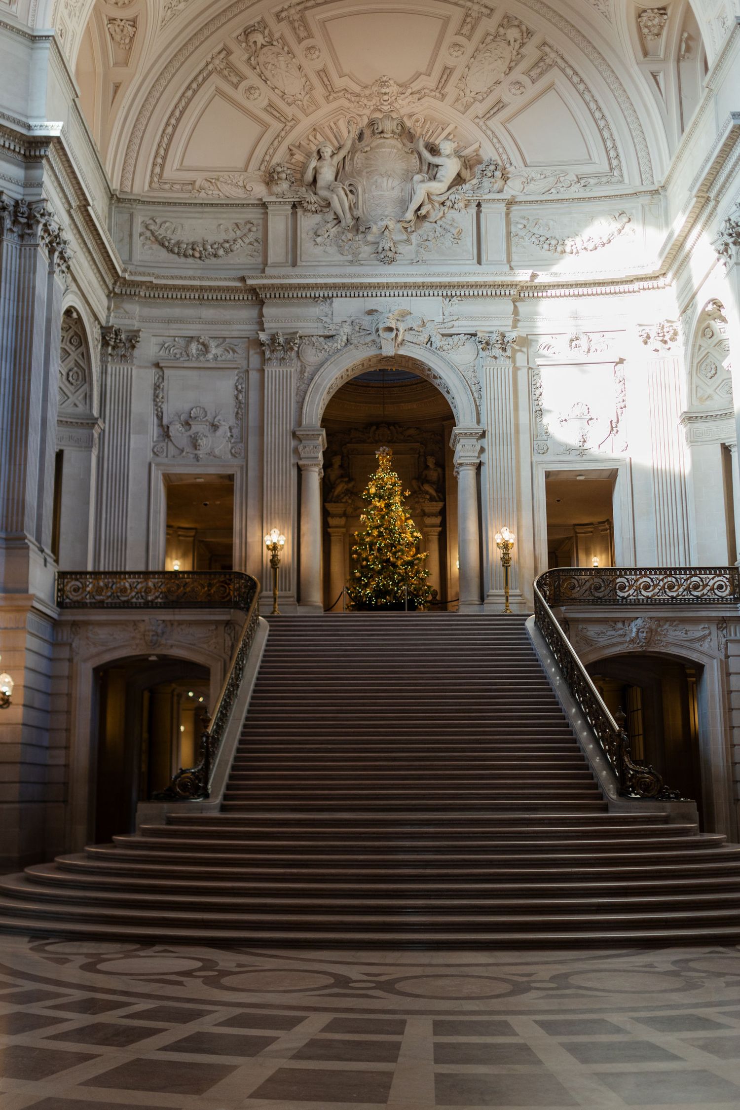 Grand staircase in San Francisco City Hall adorned with a decorated Christmas tree, ornate Beaux-Arts architecture.