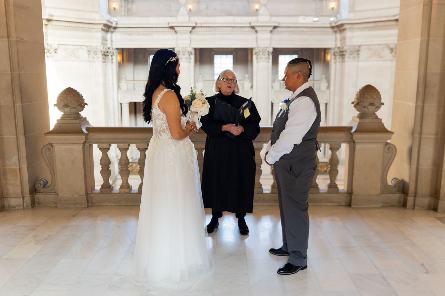 A couple exchanges wedding vows before an officiant inside an elegant marble building with ornate balustrades.