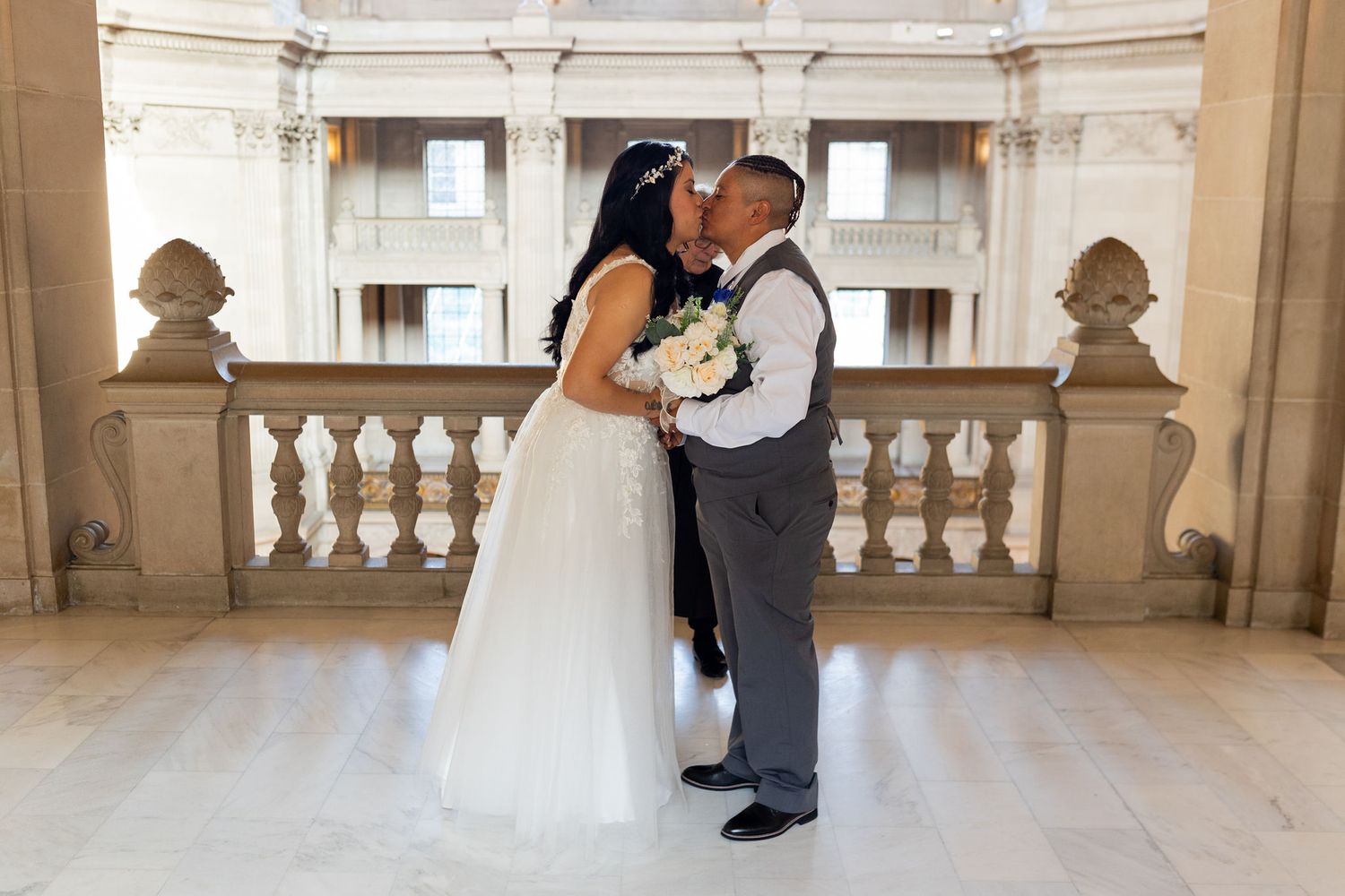 Newlywed couple sharing a romantic kiss in an elegant marble hall, bride in white gown holding bouquet, groom in gray suit.
