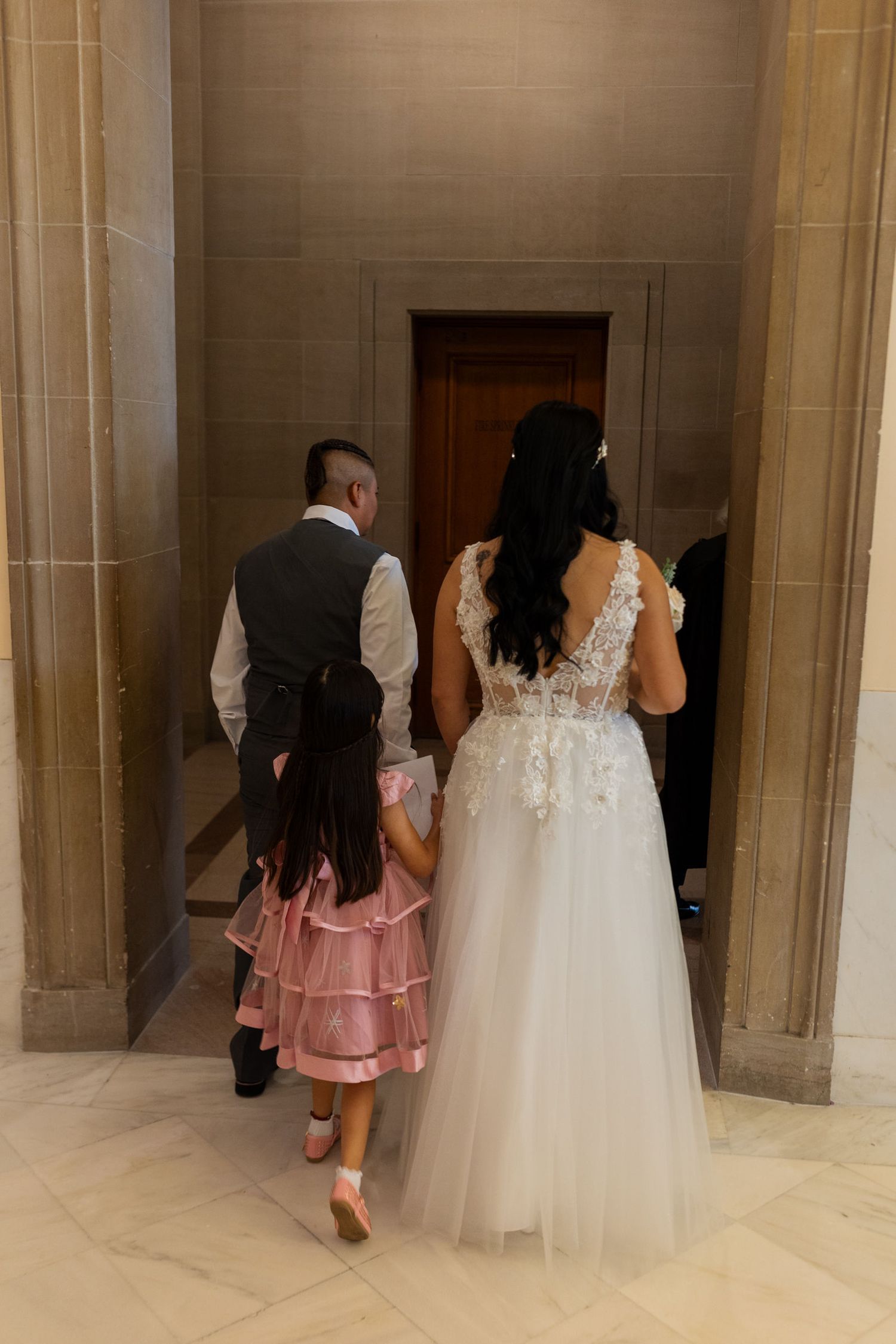 Bride in white lace gown and groom in vest walk with flower girl in pink dress through elegant marble hallway.