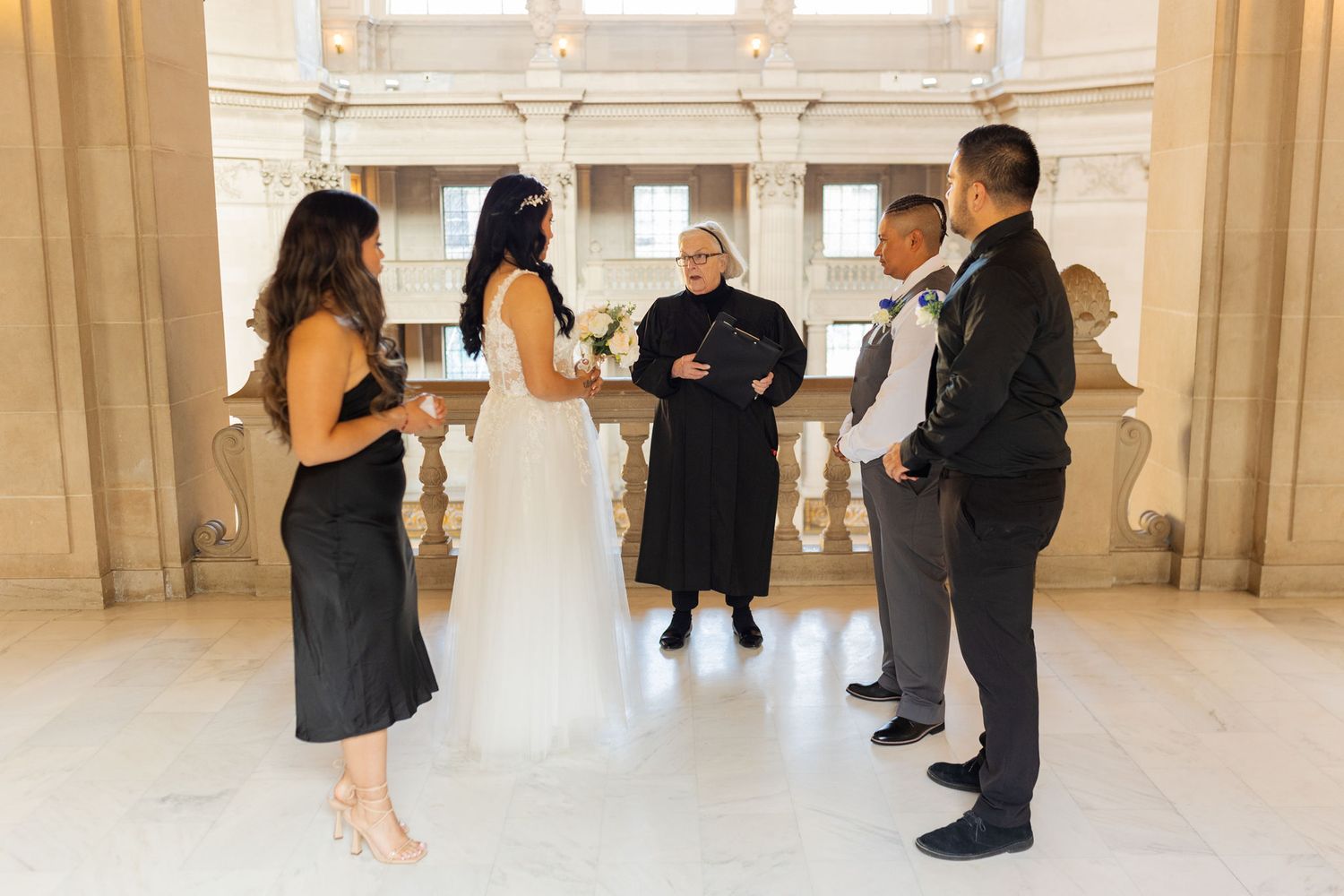 Small wedding ceremony with masked couple, officiant, and two guests in an elegant marble hall.