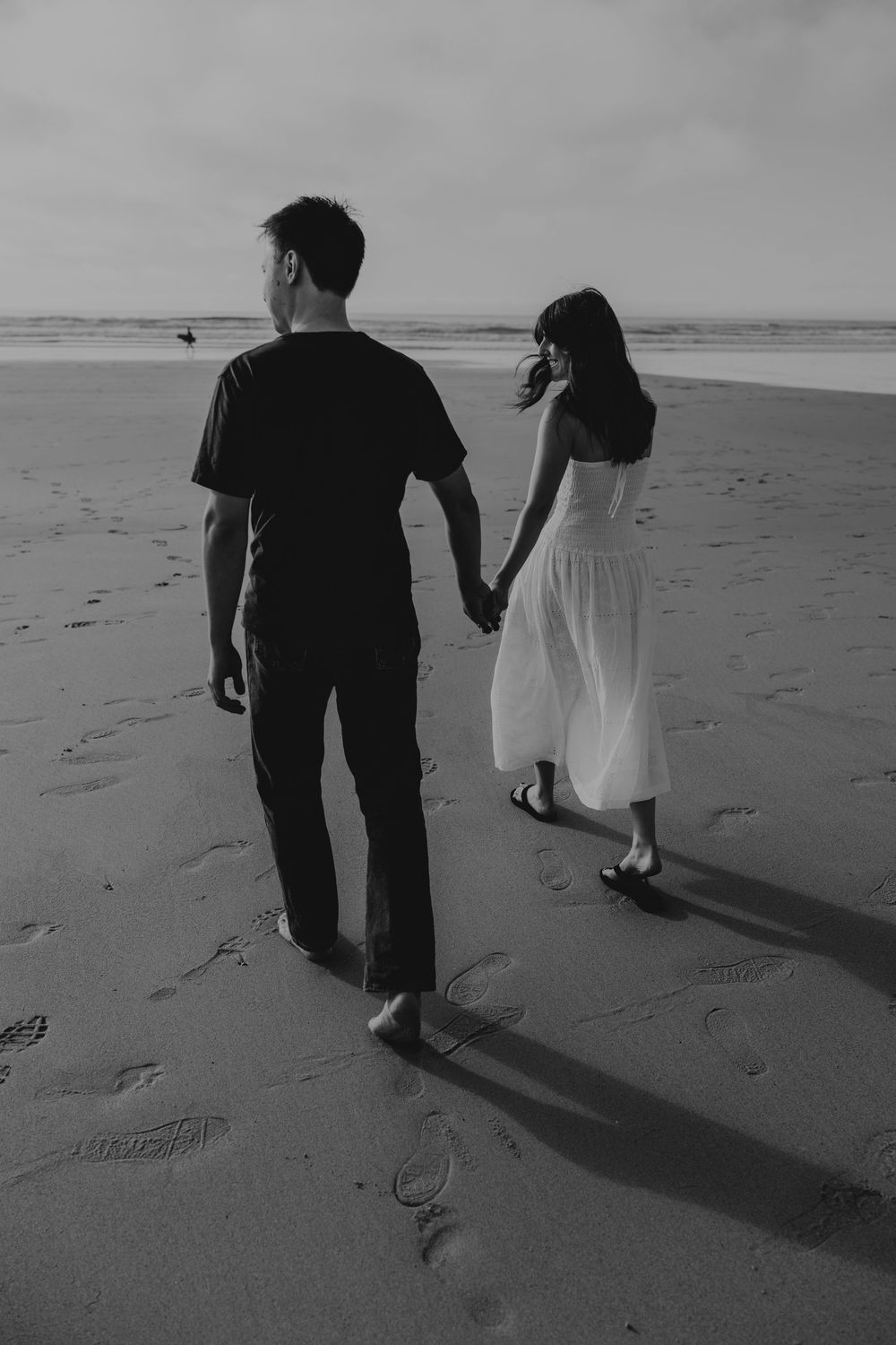 Black and white photo of a couple holding hands walking on a sandy beach leaving footprints.