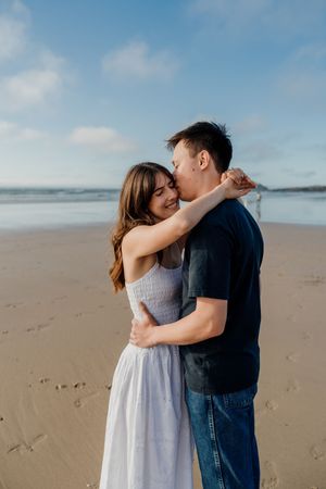A couple embraces romantically on a sandy beach, the woman in a white dress smiling as the man kisses her cheek.