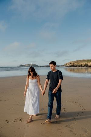 A couple holds hands while walking barefoot on a sandy beach, woman in white dress and man in black shirt and jeans.