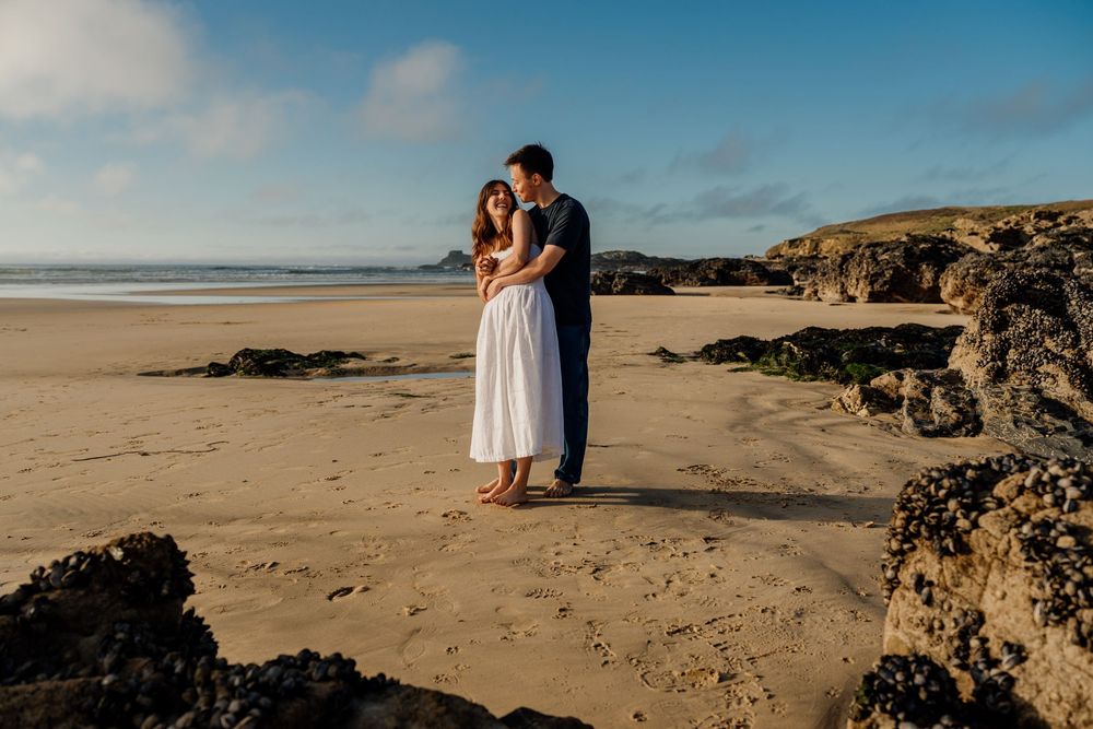 Couple embracing on a sandy beach at golden hour with rocky coastline and blue sky backdrop.
