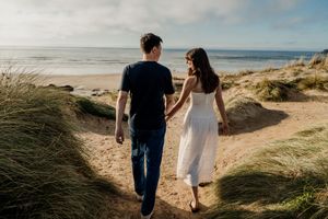 Couple holding hands walking toward a scenic beach through sand dunes with ocean views and coastal grass.