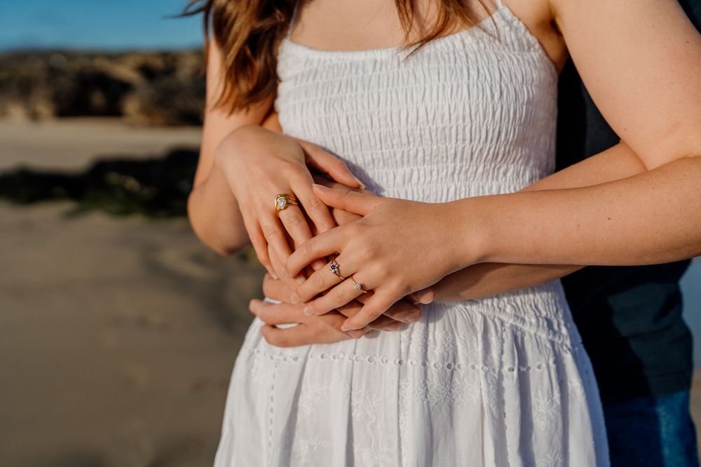 Couple showing engagement rings while embracing on a beach, woman wearing white smocked dress.
