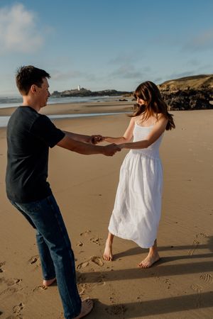 Couple holding hands and dancing barefoot on a sandy beach at golden hour, woman in white dress, man in black shirt.