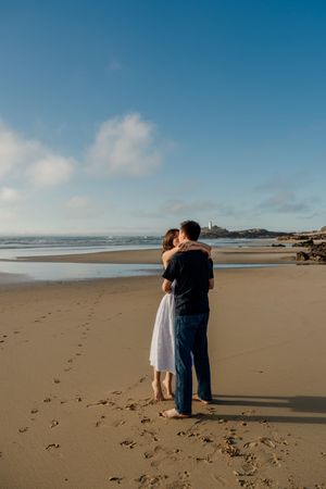 Couple embracing on a sandy beach with footprints, ocean waves, and a lighthouse in the background under a blue sky.