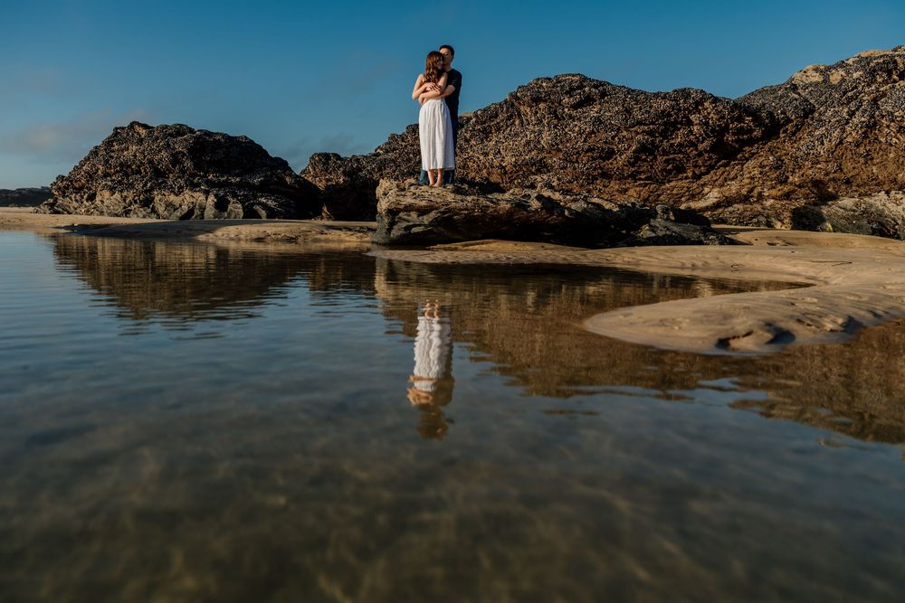 Couple embracing on rocky beach with stunning water reflection under clear blue sky.