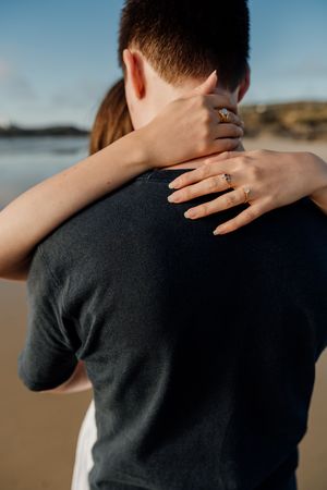 Couple embracing on a beach, woman's hand showing an engagement ring on man's shoulder.