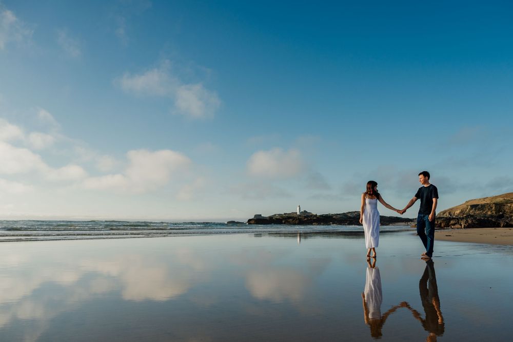 Couple holding hands on a reflective beach with a lighthouse in the background under blue sky.