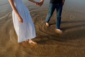 Couple holding hands barefoot on wet sandy beach, woman in white dress and man in jeans, golden hour light.