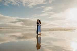 Couple kissing on a reflective wet beach at golden hour with dramatic clouds and ocean in the background.
