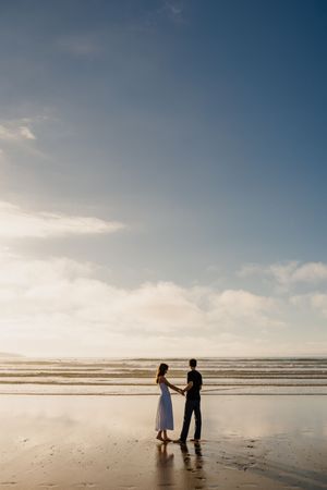 Couple holding hands on a reflective beach at sunset, woman in white dress, man in dark clothes, ocean waves behind.
