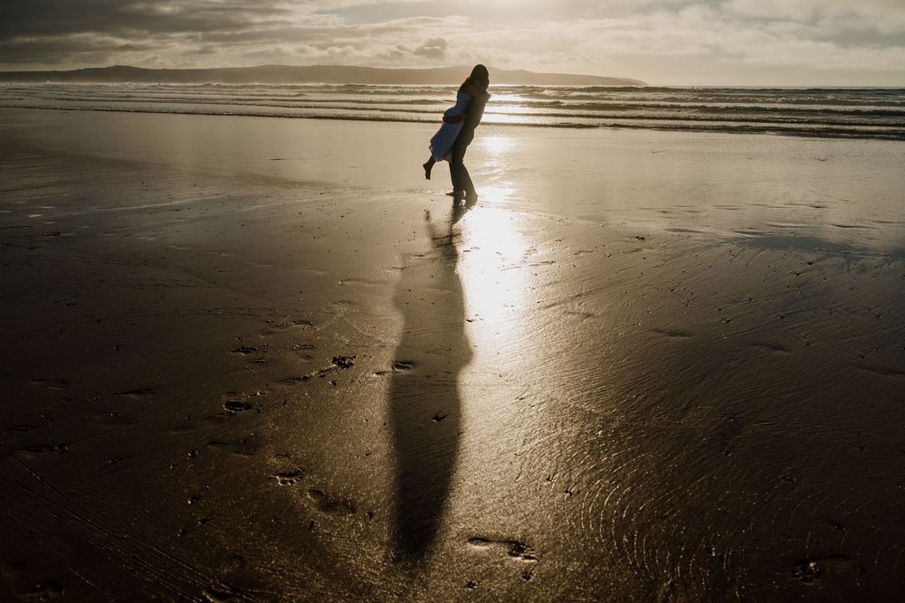 Silhouette of a person walking on a wet reflective beach at sunset with a long shadow cast on the sand.
