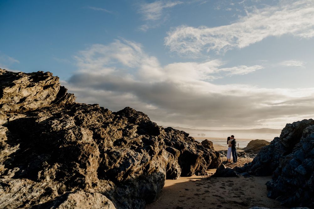 Couple embracing among rocky coastal formations with dramatic cloudy sky at golden hour.