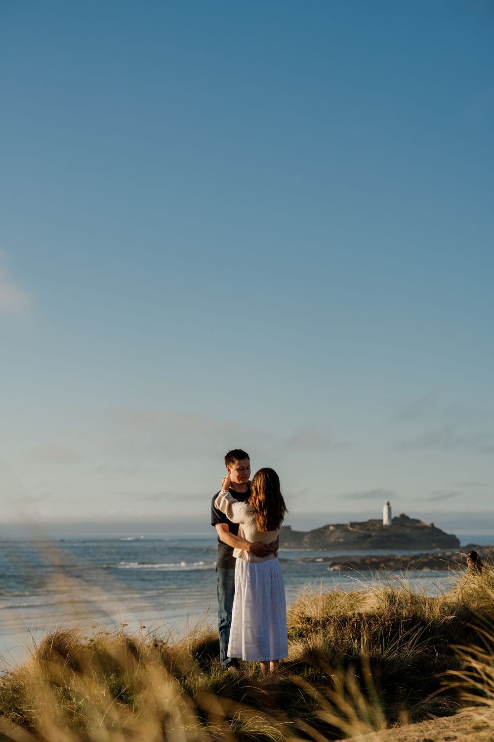 Couple embracing on coastal sand dunes with a lighthouse and ocean in the background under a clear blue sky.