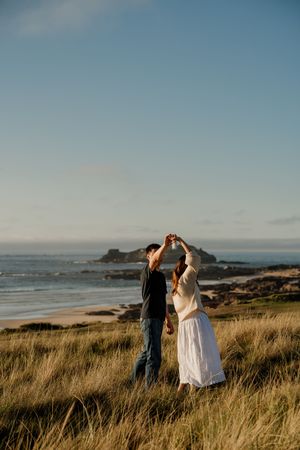 Couple dancing together in tall grass near a scenic beach at golden hour, with ocean waves and rocky shore in background.