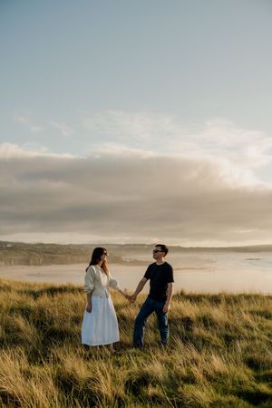 Couple holding hands in golden grass field with misty beach coastline backdrop during sunset.