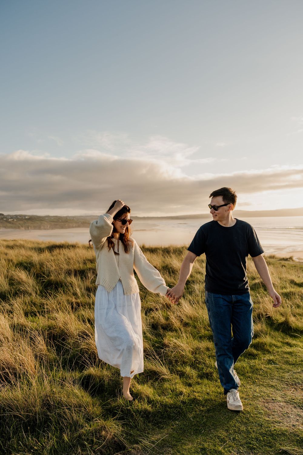 Couple holding hands walking through coastal grass dunes at golden hour with ocean backdrop.