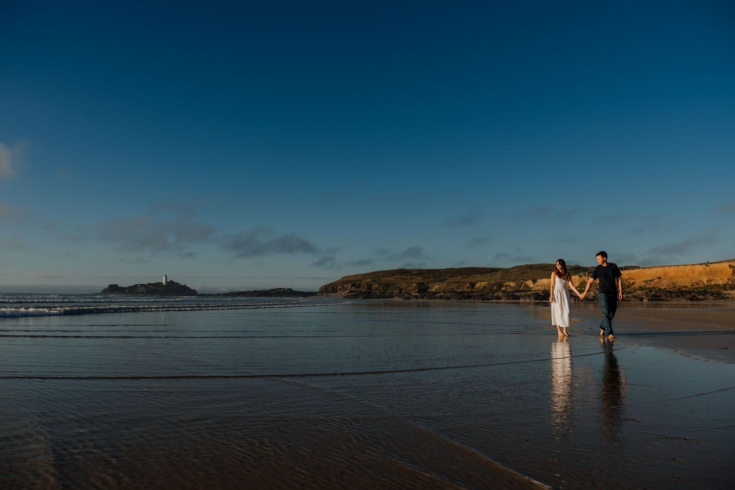 Couple embracing on a sandy beach with rocky shores and blue skies in a romantic coastal setting.