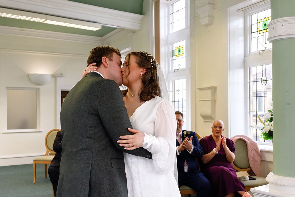 Newlyweds share first kiss at wedding ceremony while guests clap in elegant venue.