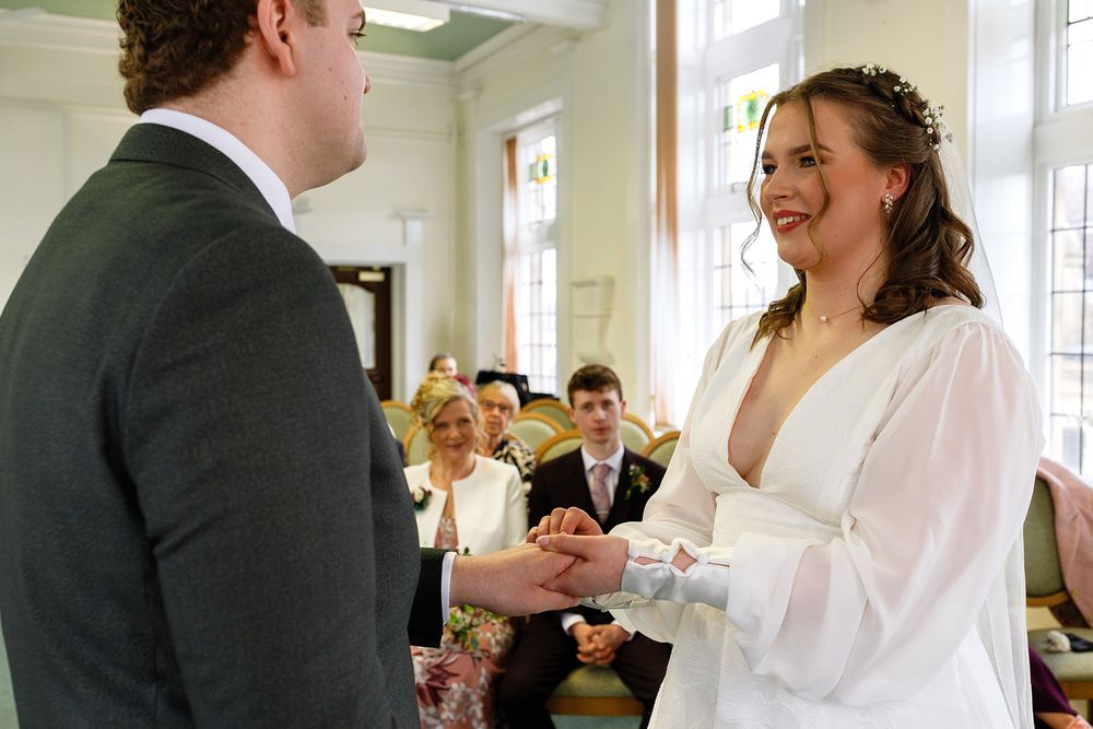 Bride and groom exchanging rings at a civil wedding ceremony, smiling joyfully, with guests seated in the background.
