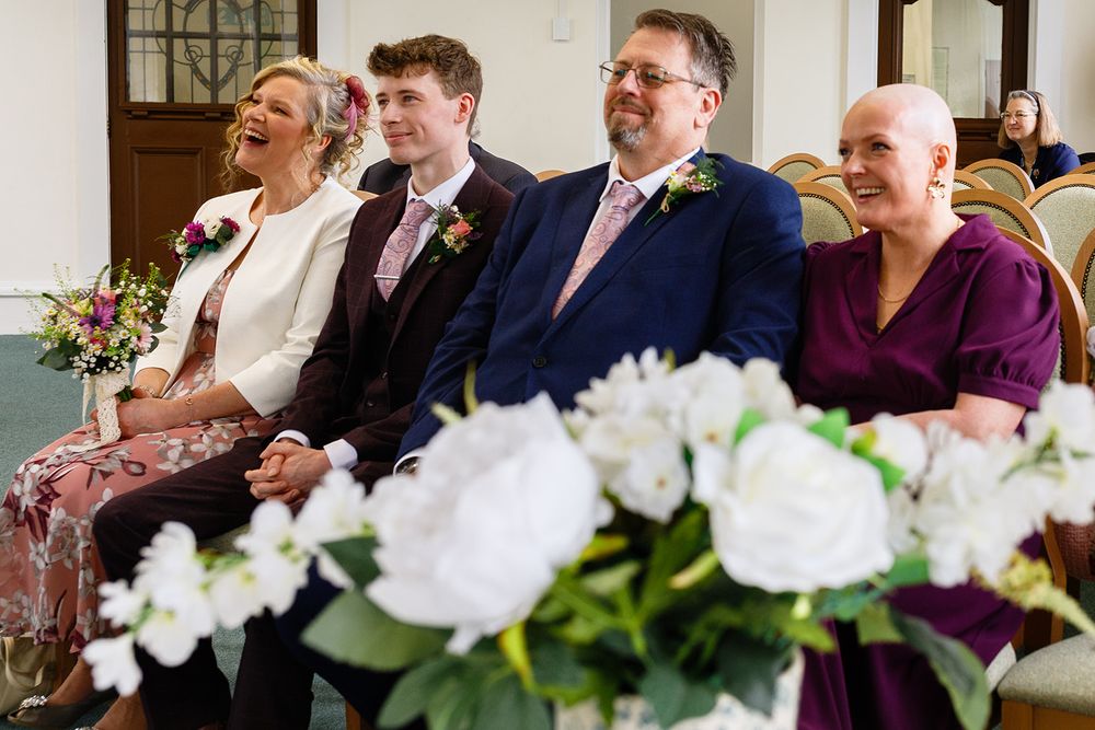 Four smiling guests seated at a wedding ceremony with white floral arrangements in the foreground.
