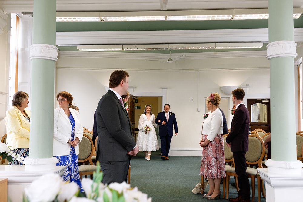 Guests gather in an elegant hall as a bride walks down the aisle during an intimate wedding ceremony.