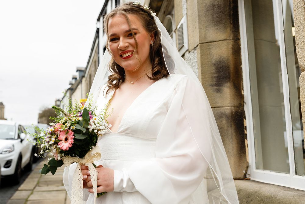 Smiling bride in white wedding dress and veil holding pink floral bouquet standing on a street.