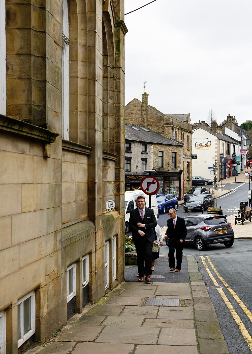 Two professionals walking on a historic stone-paved street in a quaint British town with shops and parked cars.