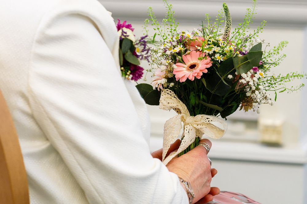 Bride in white dress holding a colorful wedding bouquet with pink gerbera daisies and wildflowers tied with a gold ribbon.