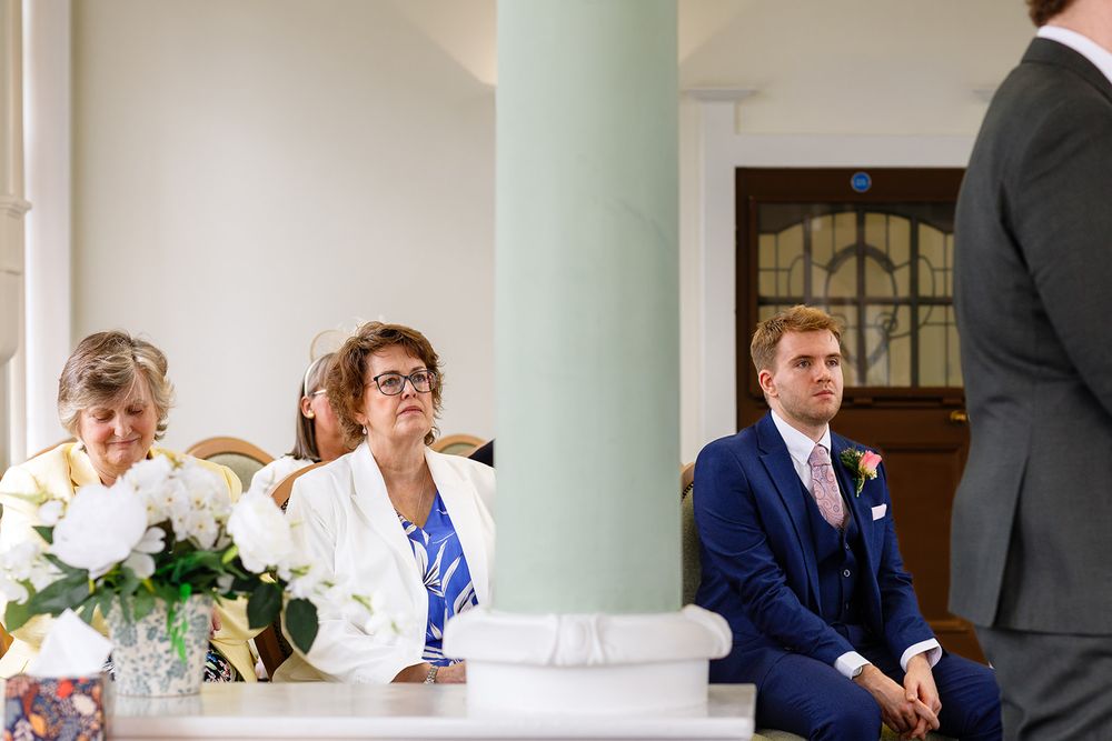 Wedding guests seated at ceremony, woman in white jacket and man in navy suit listening attentively near elegant columns.