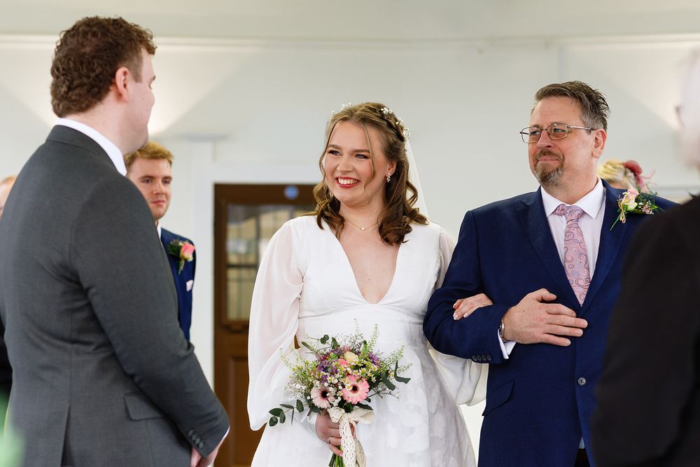 Bride in white dress holding bouquet smiles with groom and guests during indoor wedding ceremony.