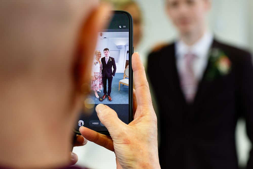 Person photographing a groom in a suit on a smartphone at a wedding venue.