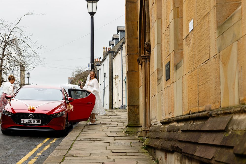 A bride in a white gown stands beside a red Mazda car on a cobblestone street next to a sandstone building.