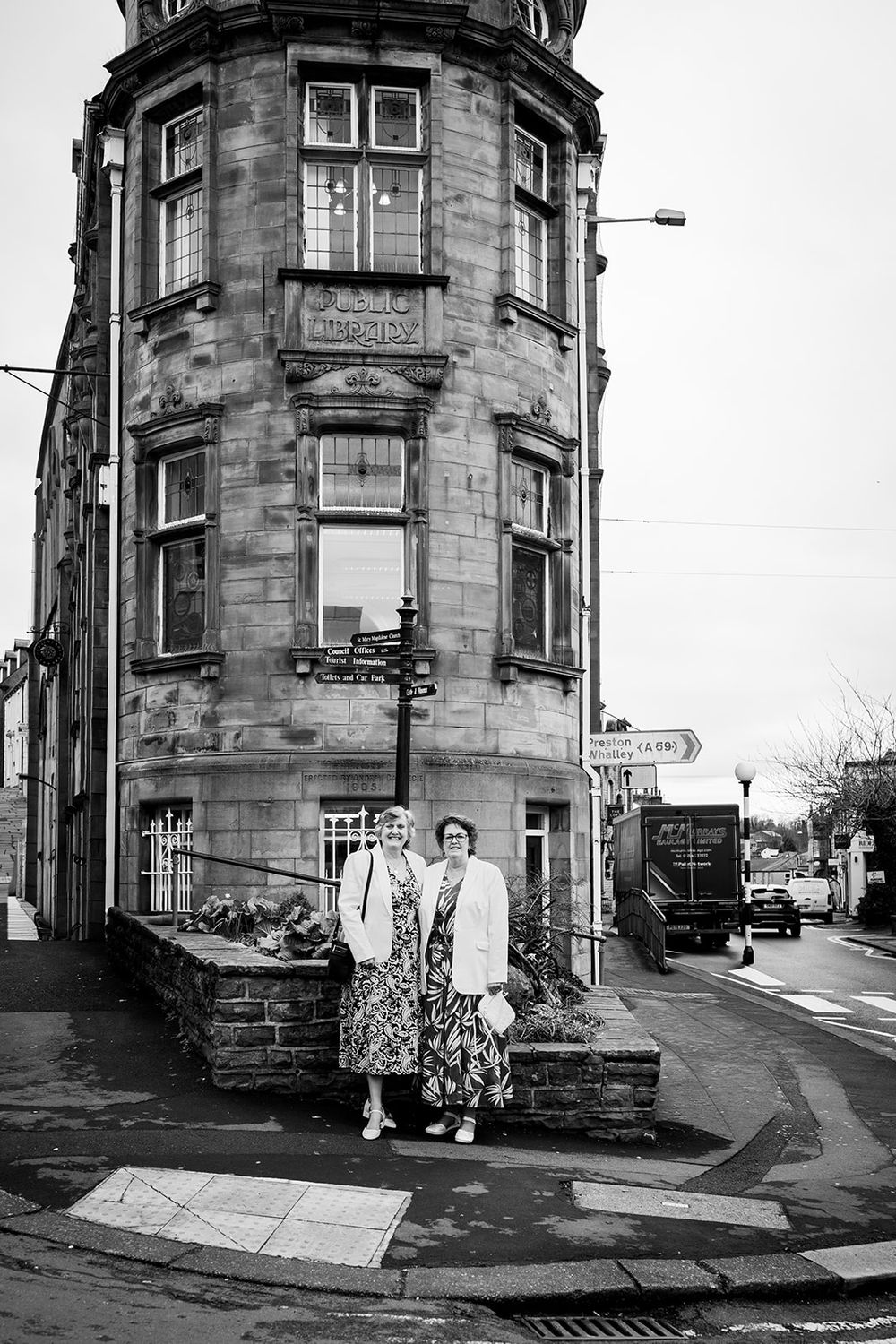 Two women posing in front of a historic stone corner building on a quiet street, black and white photography.