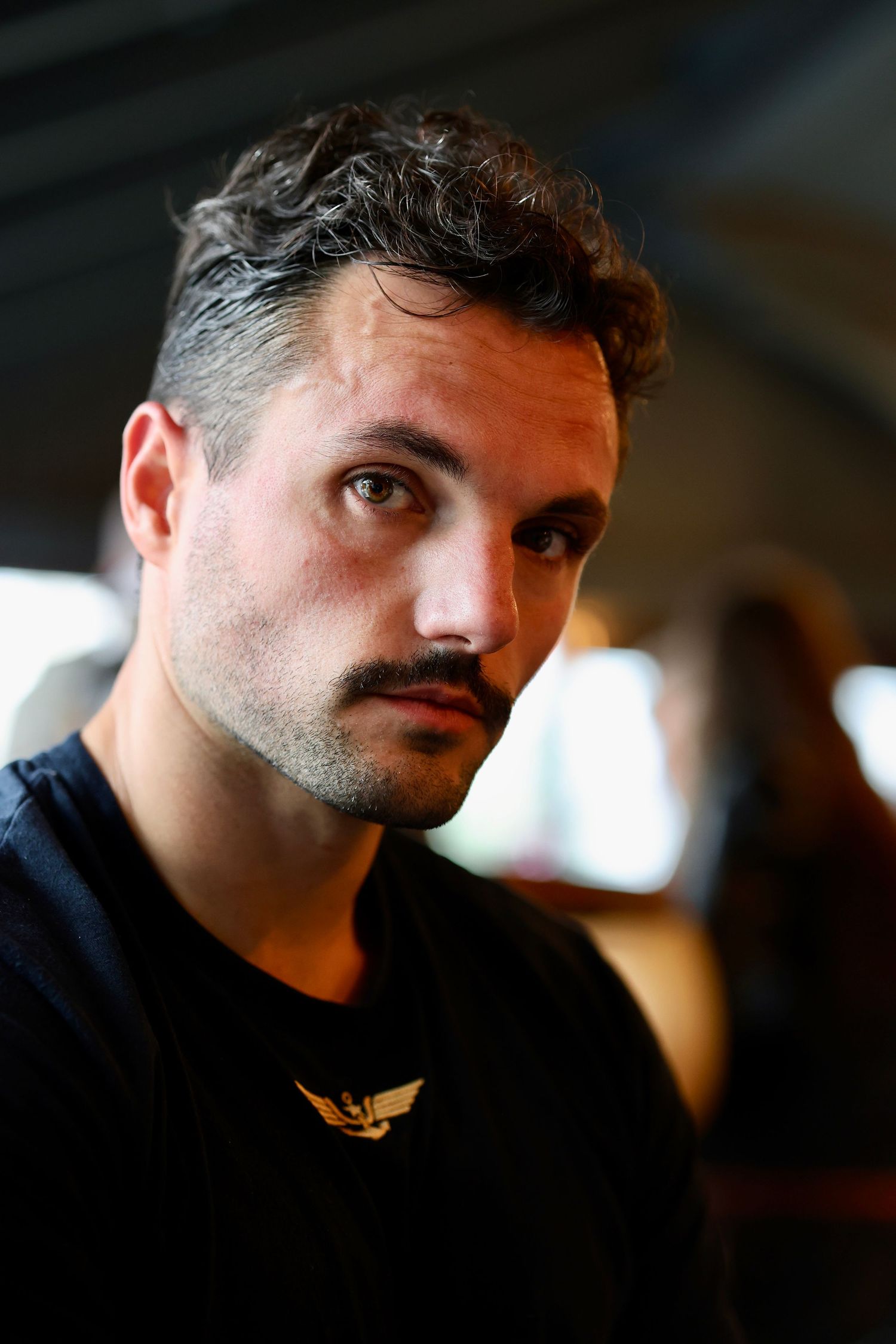 Close-up portrait of a young man with curly hair and mustache wearing a black t-shirt with gold logo.
