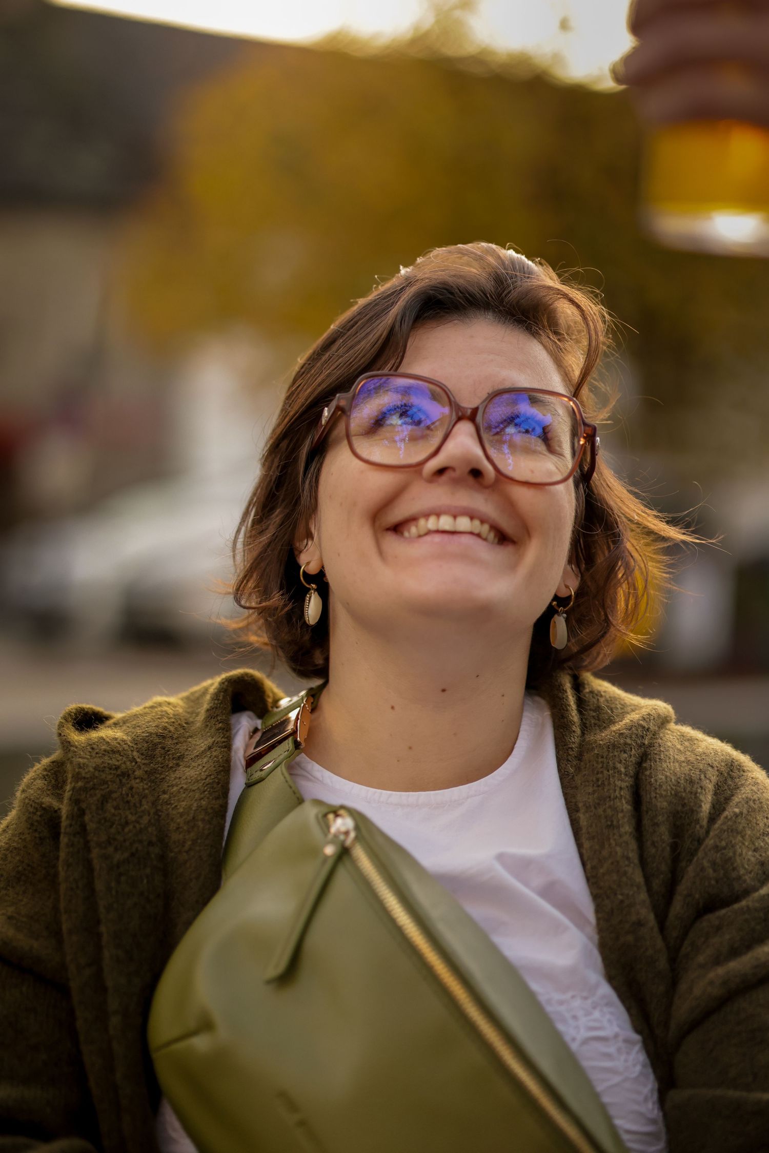 Smiling woman with glasses and short brown hair wearing a green coat and olive fanny pack outdoors in autumn.