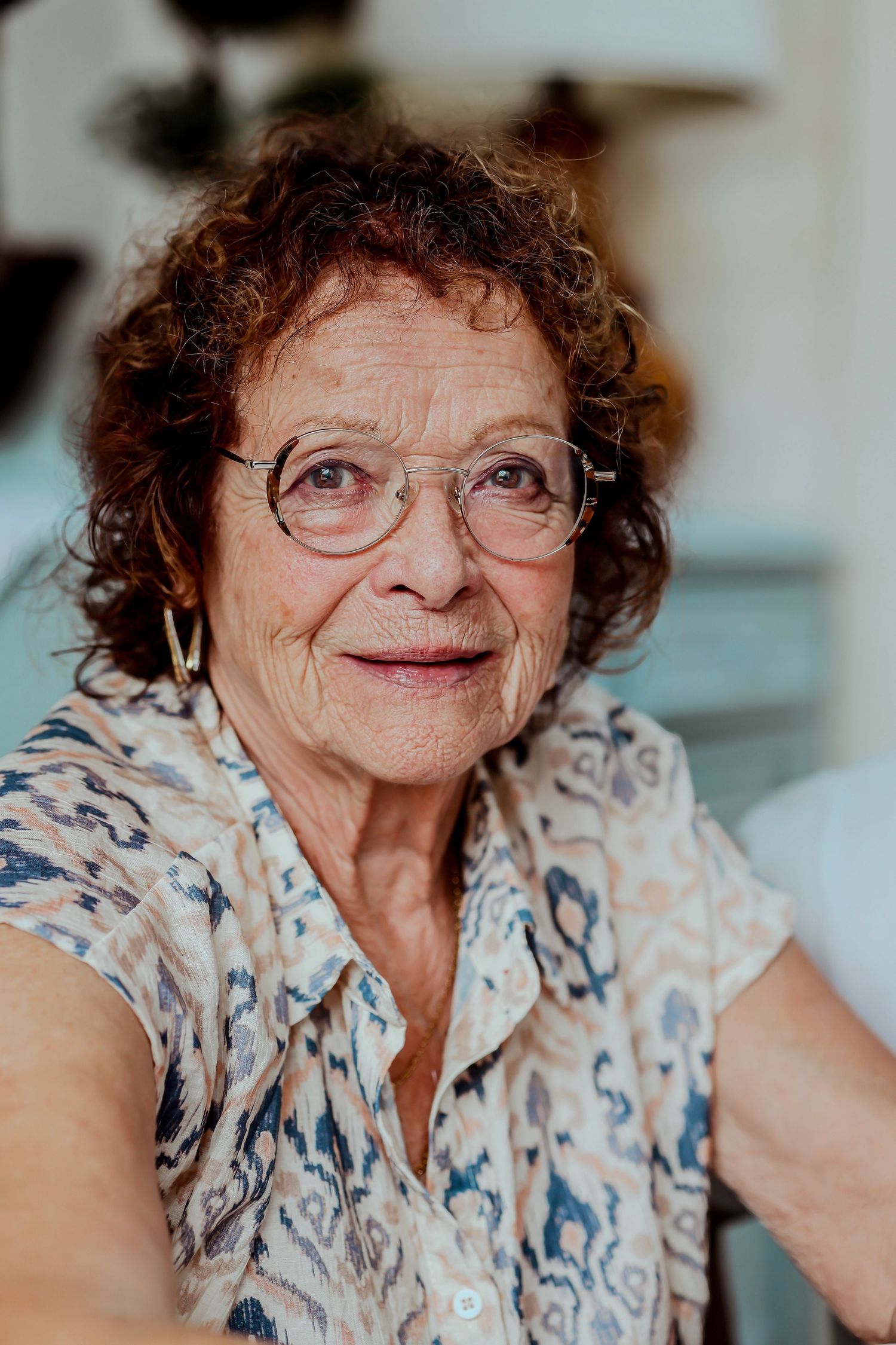 Elderly woman with curly red hair and glasses wearing a floral blouse, smiling warmly indoors.