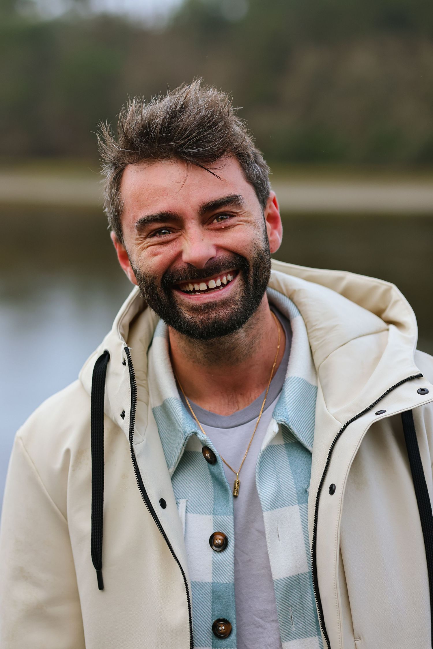 Smiling bearded man in a cream jacket and plaid shirt outdoors near a lake.