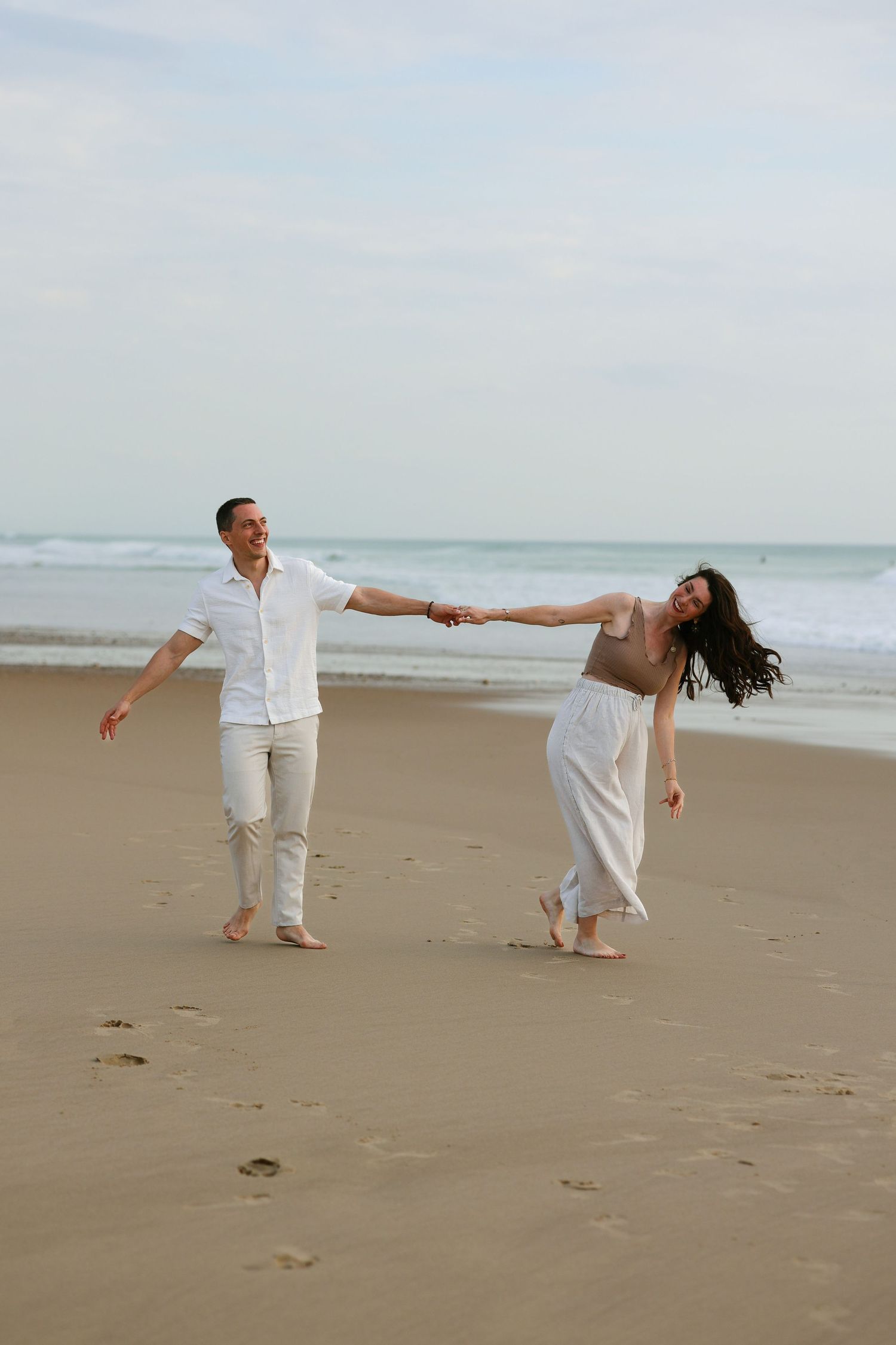 Happy couple holding hands and dancing barefoot on sandy beach with ocean waves.