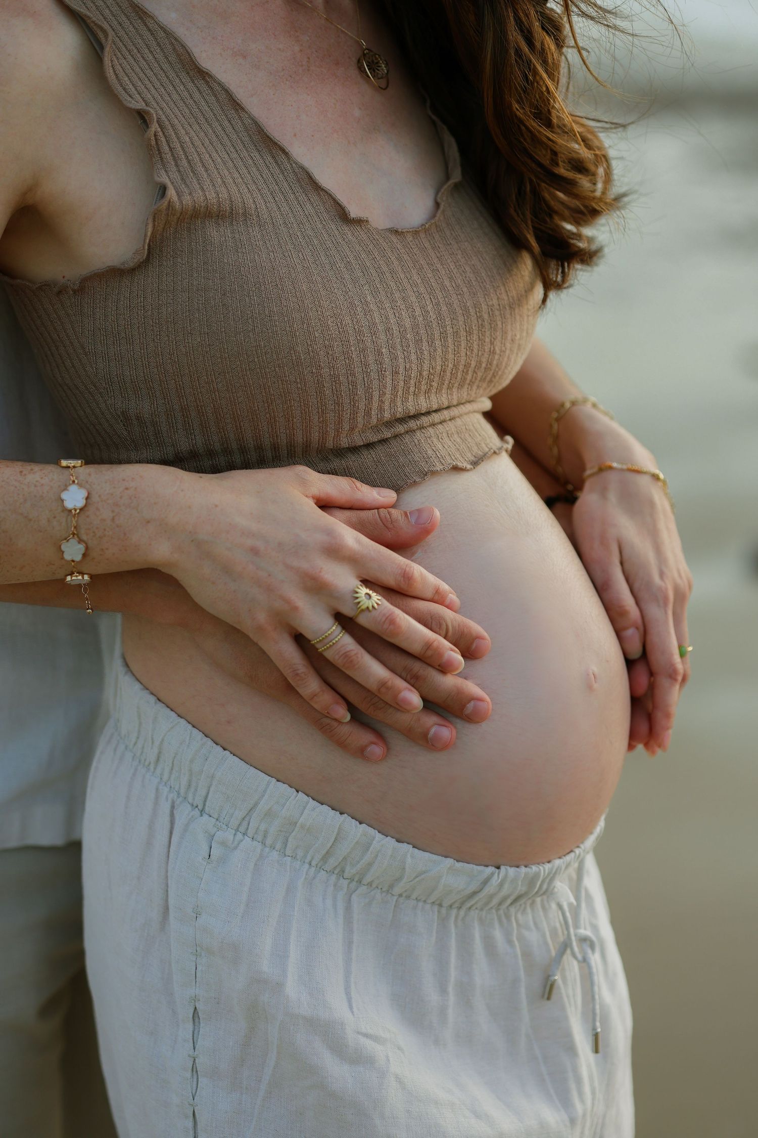Pregnant woman in tan crop top holding baby bump with partner's hands on beach.