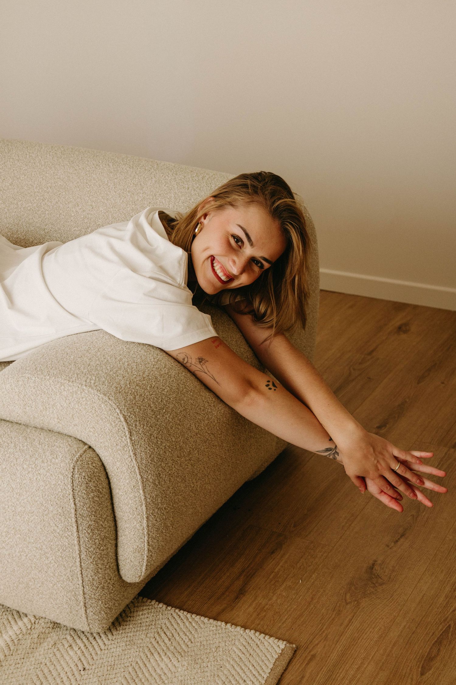 Smiling blonde woman leaning over beige sofa arm onto hardwood floor.