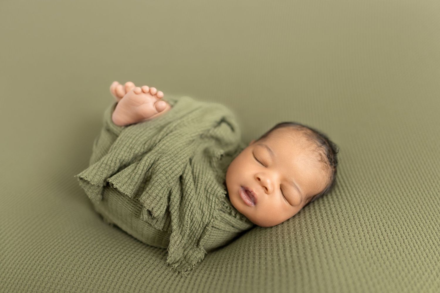 Newborn baby swaddled in olive green wrap sleeping peacefully on matching green textured backdrop.