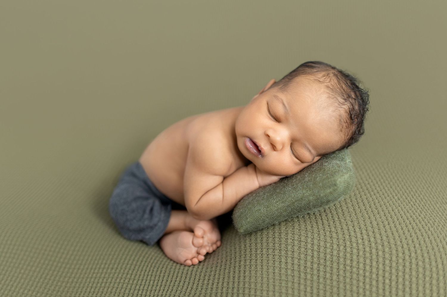 Sleeping newborn baby in blue pants posed on green pillow against olive background.